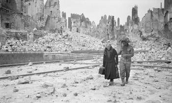 A British soldier in Caen after its liberation, gives a helping hand to an old lady amongst the scene of utter devastation.jpg